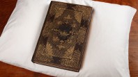 Image of a brown hard cover book with debossed gold decorations on the cover placed on a white pillow. The book is a volume of the Collected writings of King James I with personalised binding for Prince Charles, London 1616, one of the rare books in the John Emmerson Collection at State Library Victoria.