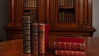 Image of rare books from the John Emmerson Collection at State Library Victoria placed on a brown table. Three books on the left are placed upright, and two on the right are stacked on top of each other. There is a glass bookcase in the background.