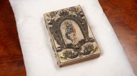 Image of a rare book with embroidered binding thought to belong to Queen Henrietta Maria, wife of King Charles I, from the John Emmerson Collection. The binding is ornate and detailed, and can be found across the entire book. The volume sits on a white pillow placed on a brown table.