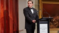A man in a suit and bow tie speaks at a podium