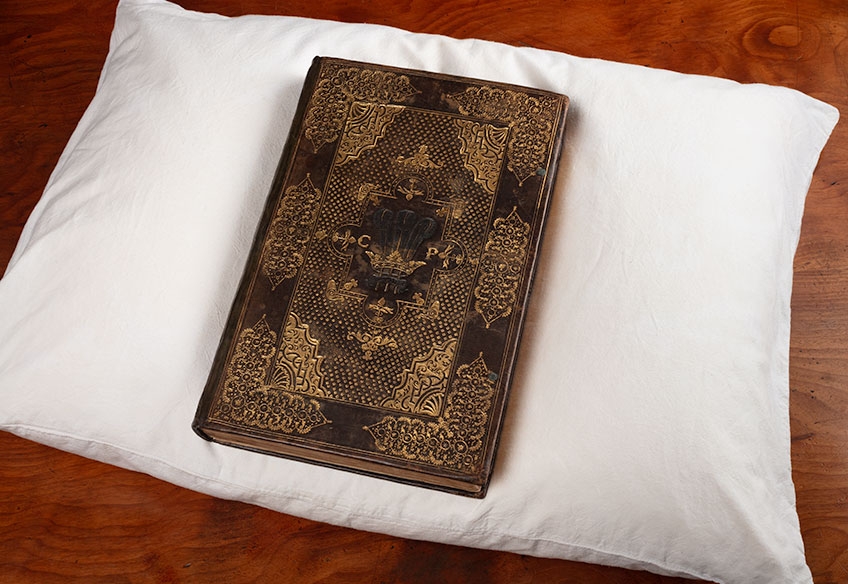 Image of a brown hard cover book with debossed gold decorations on the cover placed on a white pillow. The book is a volume of the Collected writings of King James I with personalised binding for Prince Charles, London 1616, one of the rare books in the John Emmerson Collection at State Library Victoria.