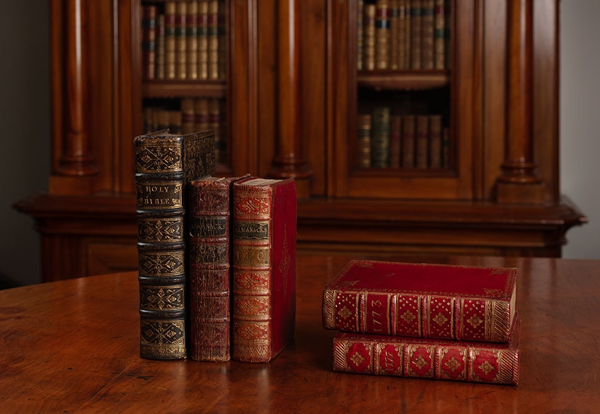 Image of rare books from the John Emmerson Collection at State Library Victoria placed on a brown table. Three books on the left are placed upright, and two on the right are stacked on top of each other. There is a glass bookcase in the background.