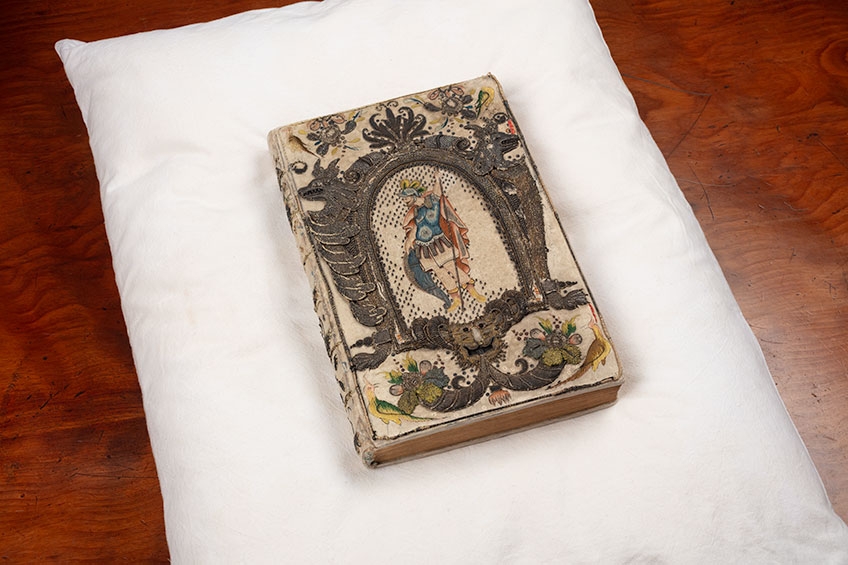 Image of a rare book with embroidered binding thought to belong to Queen Henrietta Maria, wife of King Charles I, from the John Emmerson Collection. The binding is ornate and detailed, and can be found across the entire book. The volume sits on a white pillow placed on a brown table.