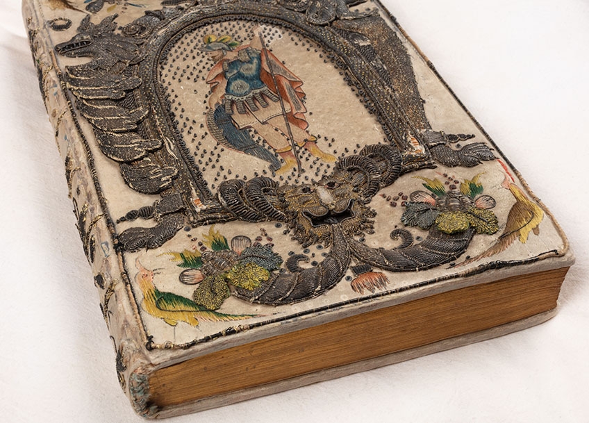 Close up image of a rare book with embroidered binding thought to belong to Queen Henrietta Maria, wife of King Charles I, from the John Emmerson Collection. The binding is ornate and detailed, and can be found across the entire book. The volume is placed on a white pillow.