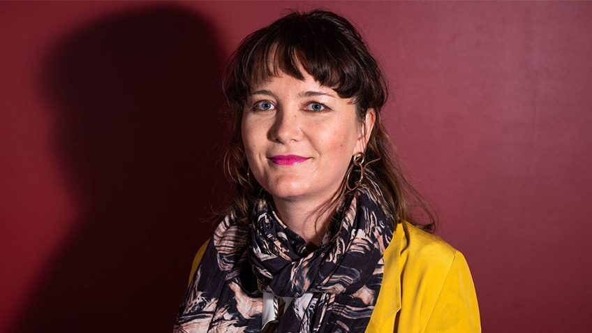 portait of woman with short hair wearing scarf and yellow jacket against red background