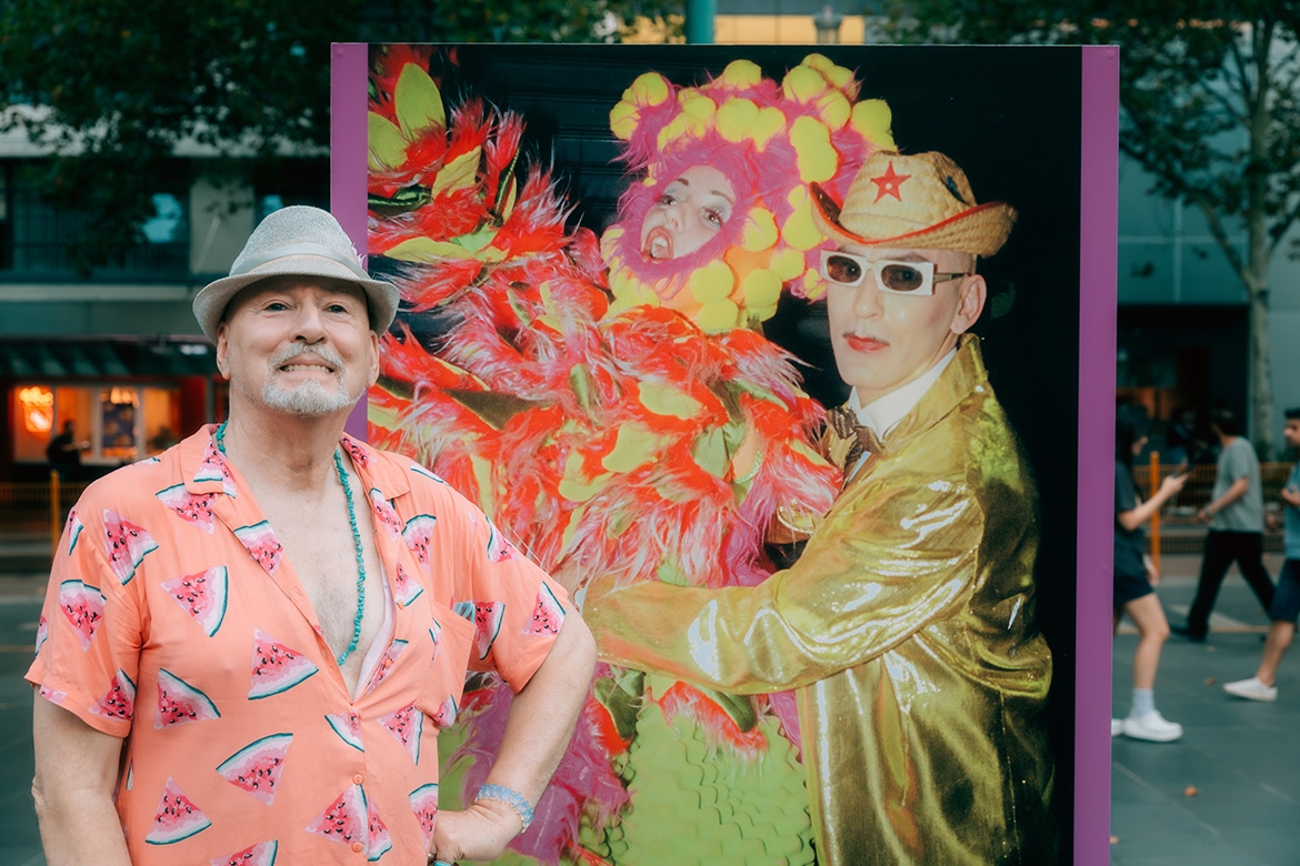 Photograph of Tim McKew standing outside State Library Victoria, in front of a photograph of himself taken by Rennie Ellis in 1978. Tim McKew is wearing a hat, beaded necklace and a peach shirt with watermelon motif. 