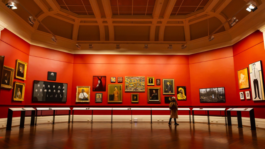 Wide angle view of the North Rotunda at State Library Victoria. The room is painted red and features artwork from the State Collection hung salon-style on its walls. There is a visitor viewing the artwork on the right side of the room.