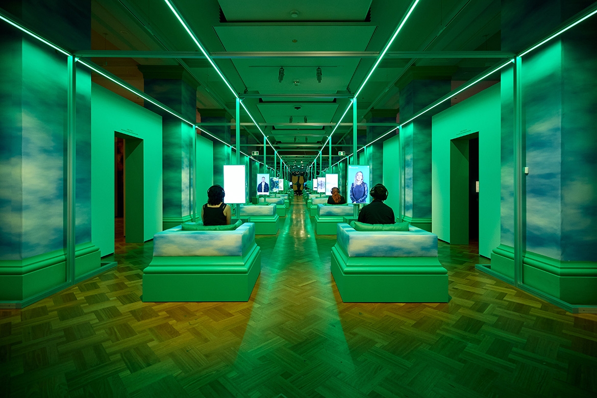 Image of an exhibition space with three people viewing video works in individual seats. The room is lit in neon green, with a mirrored back wall which creates an illusion of the room continuing infinitely.