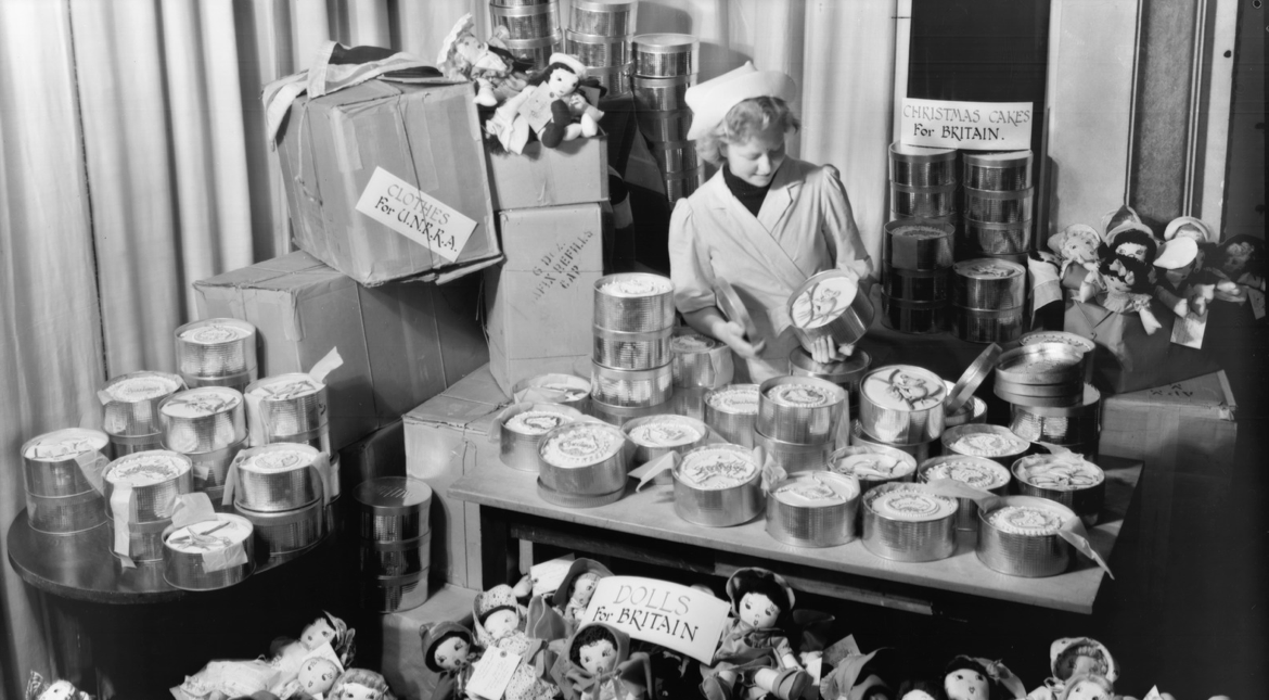 A woman in an apron and chef's hat stands behind a table piled with fresh cakes in baking tins