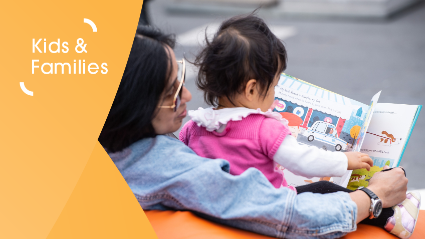 A baby being held by her mother whilst holding a picture book and turning the pages.