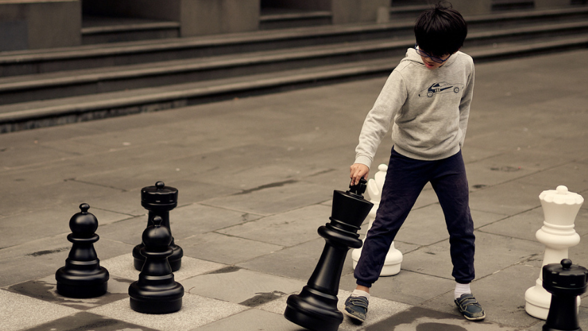 A boy in a grey hoodie and blue trackpants playing chess in the Library's forecourt.