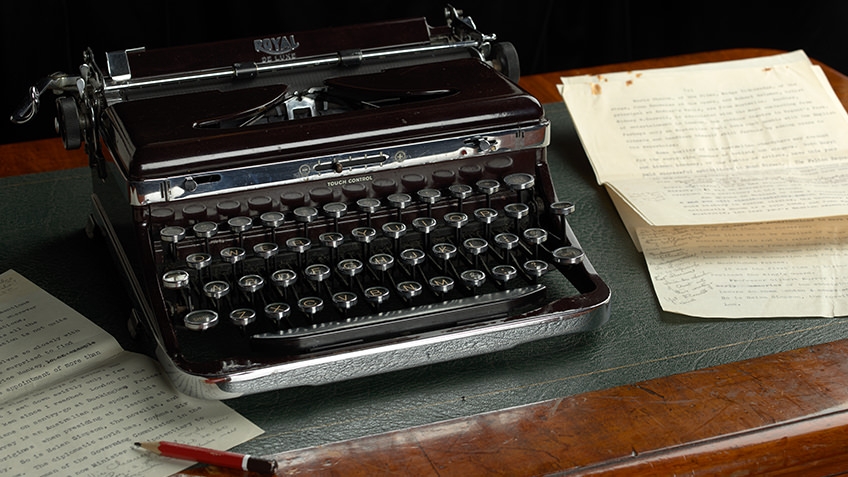 A manual typewriter sits beside typed pages on a leather-padded desk