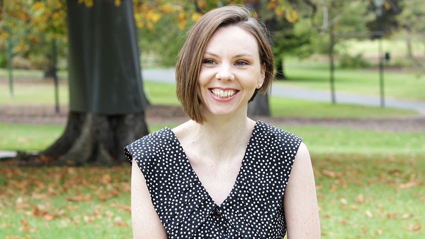 Woman standing in parkland in front of autumnal tree, wearing v-neck black dress with white dots, smiling