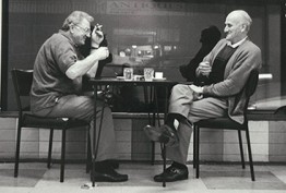 A black and white image of two men sitting outside a coffee shop sipping drinks and smoking