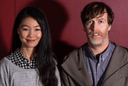 portrait of smiling woman with long dark hair and man with beard against red background