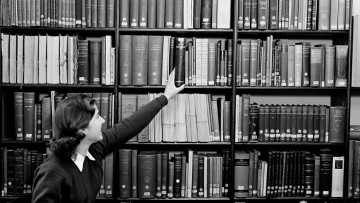 A woman reaches up to a bookshelf to take hold of a book