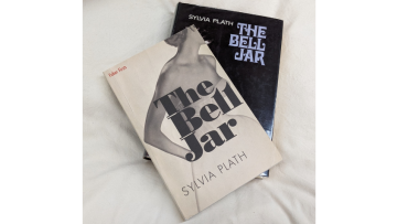 Photograph of two versions of The Bell Jar by Sylvia Plath, sitting on top of each other, slightly fanned out, on top of a pillow. 
