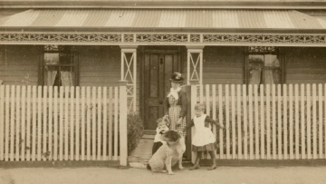 Weatherboard house with corrugated iron roof and porch, picket fence. A woman standing at gate holding the hand of a toddler, a little girl on her left, dog in front. House has a sign attached to porch roof over door 