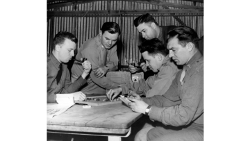 Black and white image of five officers around a table playing a board game, listening to the radio, writing letters and two are smoking pipes