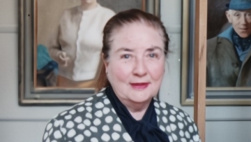 Joyce McGrath head and shoulders portrait, her self-portrait hangs on the wall in the background.
