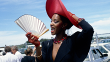 A black woman in black dress and blight red hat, smiling and waving her white fan