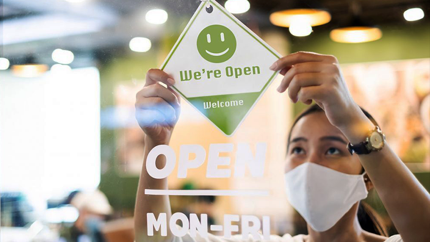 A woman wearing a face mask places a 'Welcome' sign on a shop door
