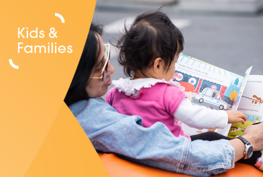 A baby being held by her mother whilst holding a picture book and turning the pages.