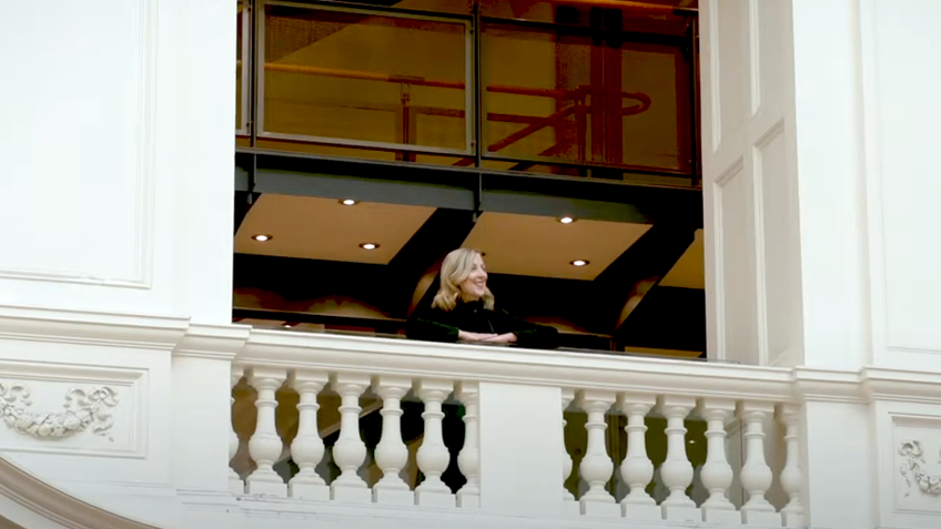 A woman in a black outfit stands on an ornate, white balcony indoors