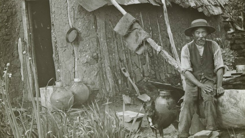 Old black and white photo of a Chinese miner sitting outside his wattle and daub hut with his dog